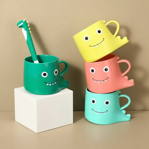 Colorful children's mugs with smiling faces, a green dinosaur-shaped toothbrush, and a box on a green background.